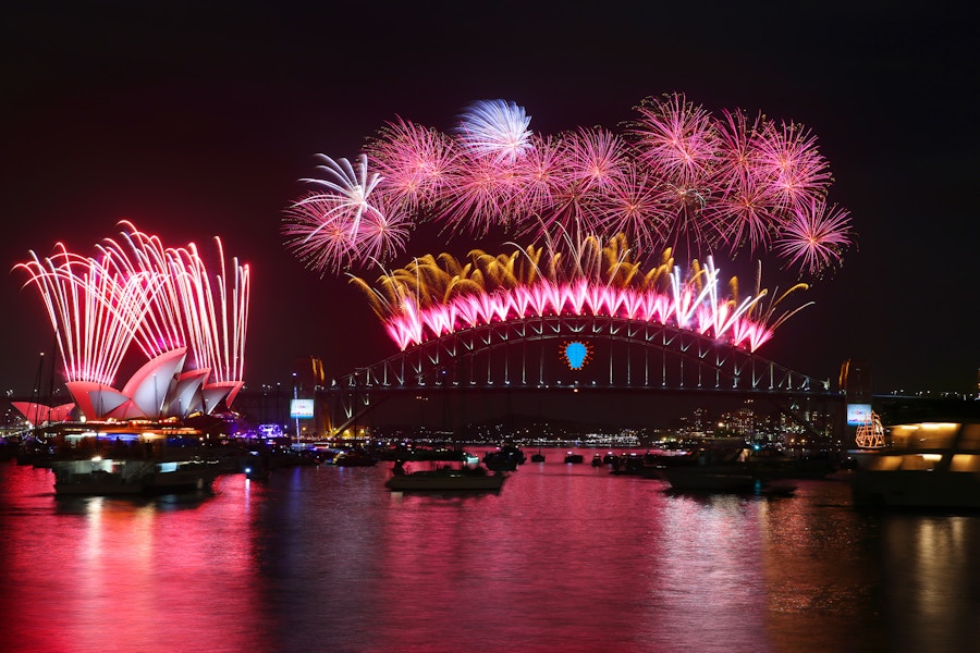 Fireworks illuminate the night sky above the Sydney Opera House and Sydney Harbour Bridge, reflecting on the water with boats in the foreground.