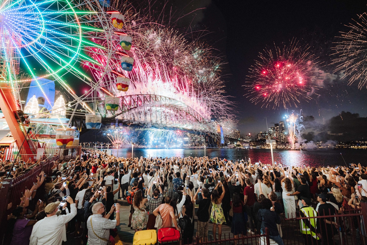 A crowd watches and records colorful fireworks exploding over a harbor with a large ferris wheel and bridge illuminated at night.