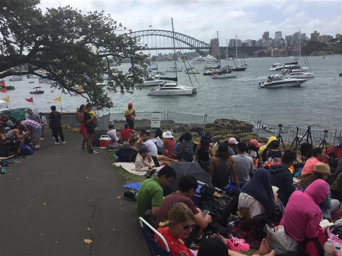 People sit and gather along a waterfront path with boats in the harbor and the Sydney Harbour Bridge visible in the background on a cloudy day.