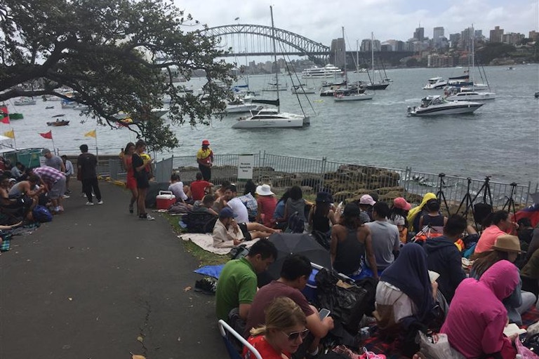 People sitting on the ground and near the water, looking towards Sydney Harbour Bridge with boats in the water under a partly cloudy sky.