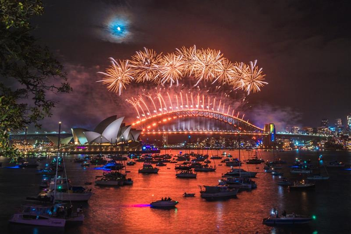 Fireworks light up the night sky above Sydney Harbour Bridge and Opera House, reflecting on the water with boats gathered below.