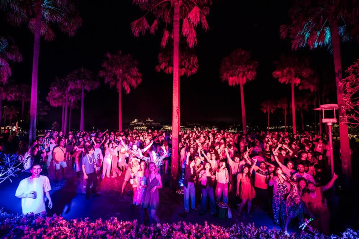 A large crowd gathers outdoors at night under palm trees, illuminated by colorful lights, with many people dancing and raising their arms.