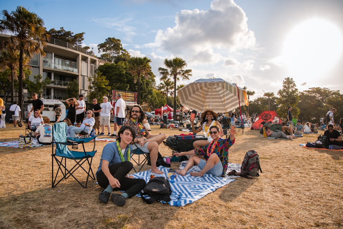 A group of people sits on blankets and chairs at an outdoor event, surrounded by others on a grassy field with trees, buildings, and a bright sun in the background.