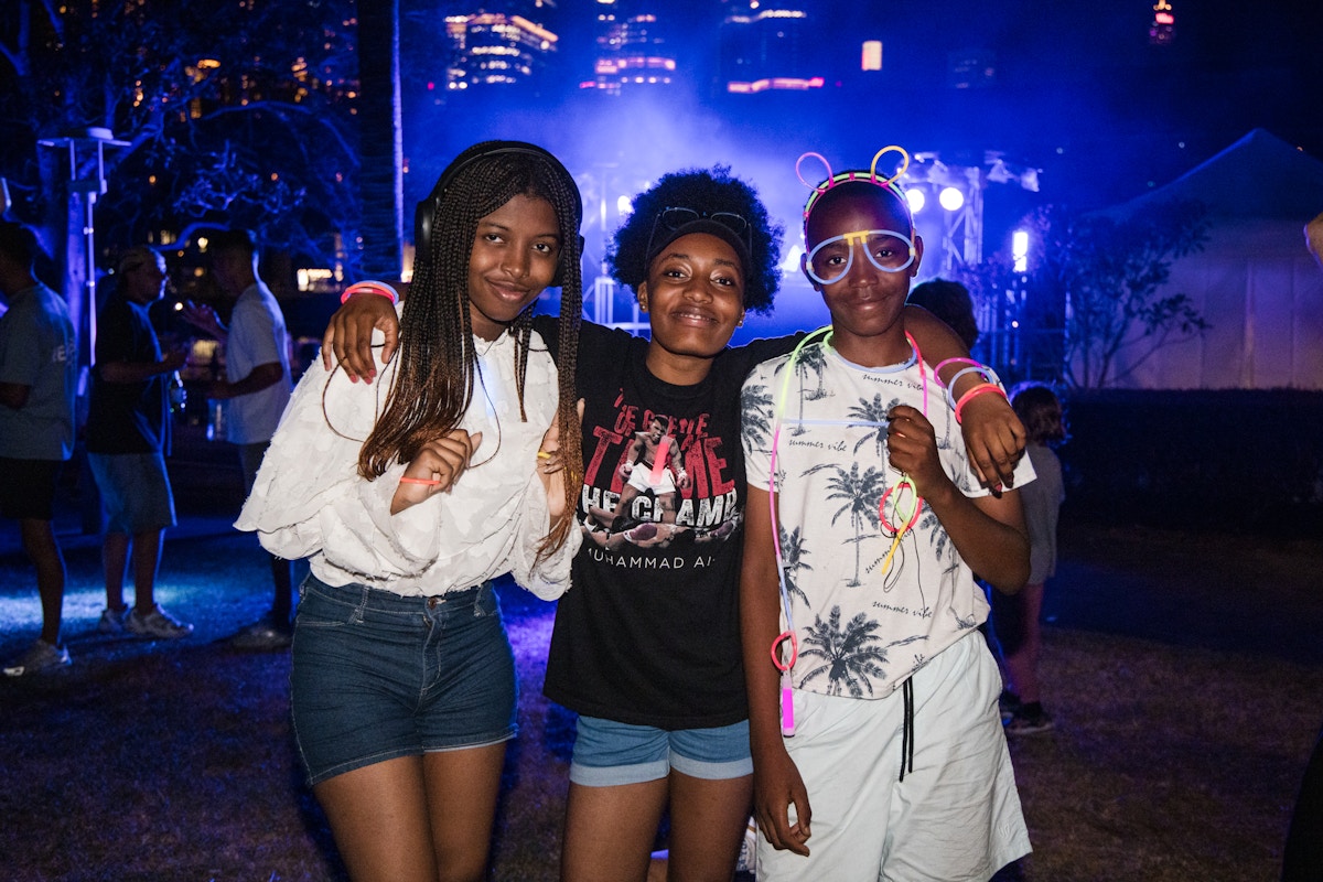 Three teens pose together at an outdoor nighttime event, dressed in casual summer clothes with light-up accessories, city lights and a stage in the background.