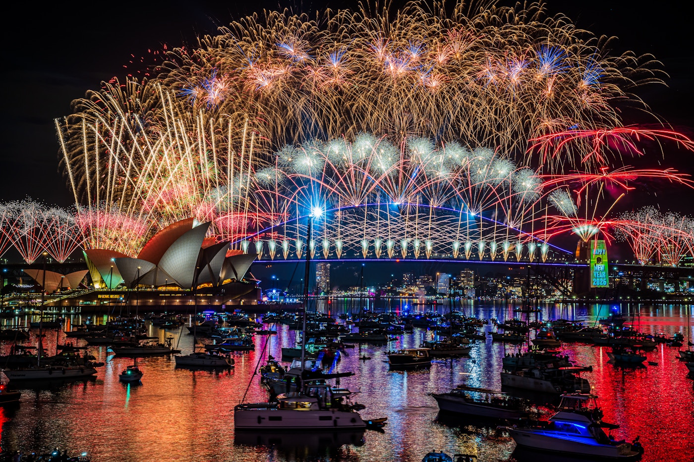 Fireworks light up the night sky over Sydney Harbour, with the Opera House and Harbour Bridge illuminated, and boats on the water reflecting the colorful display.