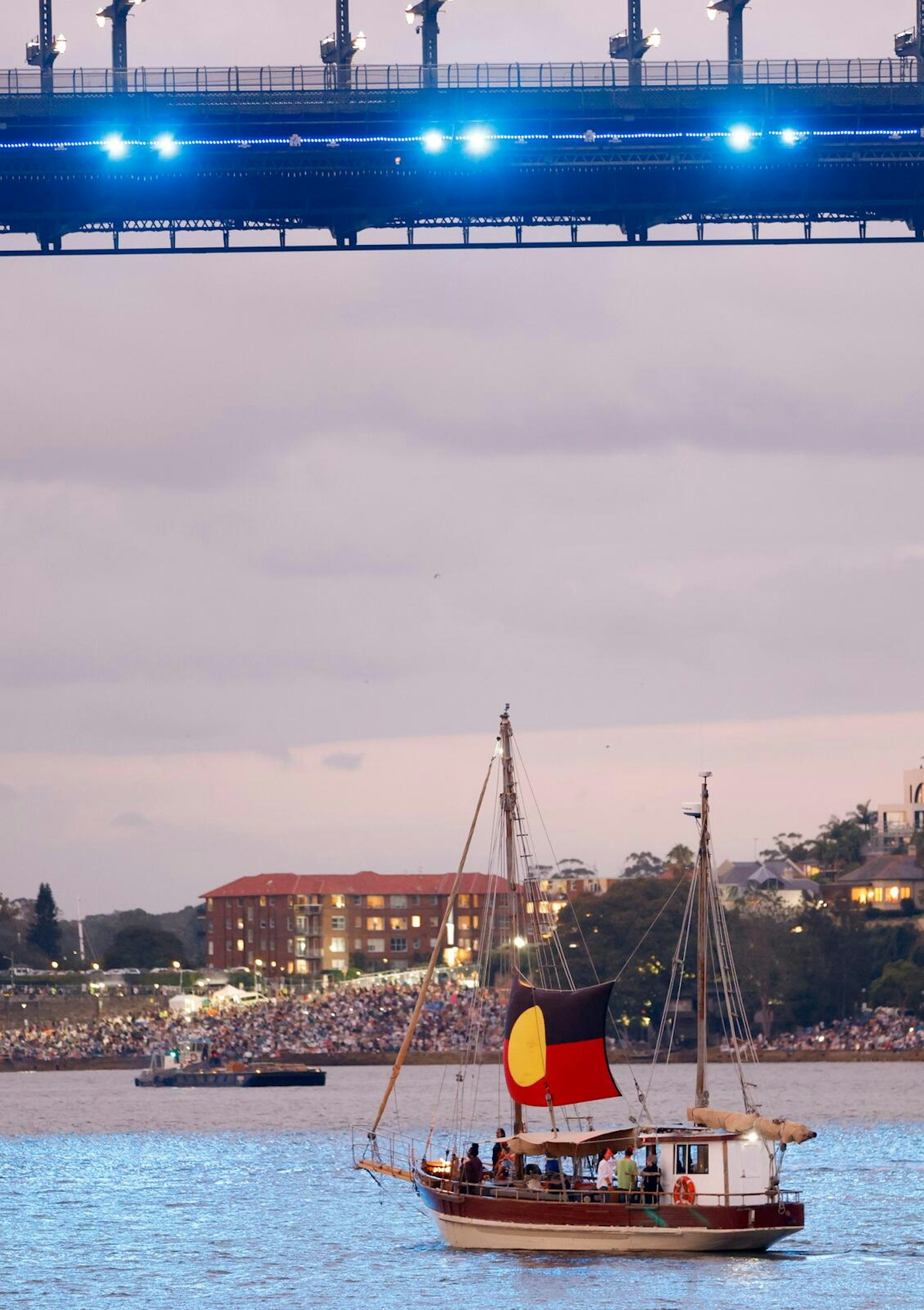 A sailboat with an Aboriginal flag sails on a river at dusk with a bridge above and a crowd gathered on the opposite bank.