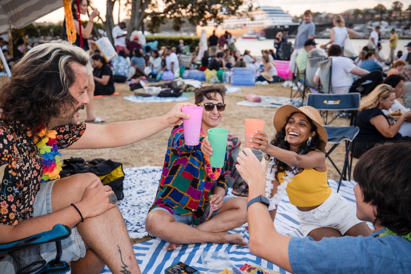Four people sitting on a blanket outdoors raise colorful cups in a toast at a picnic; a crowd and trees are visible in the background.
