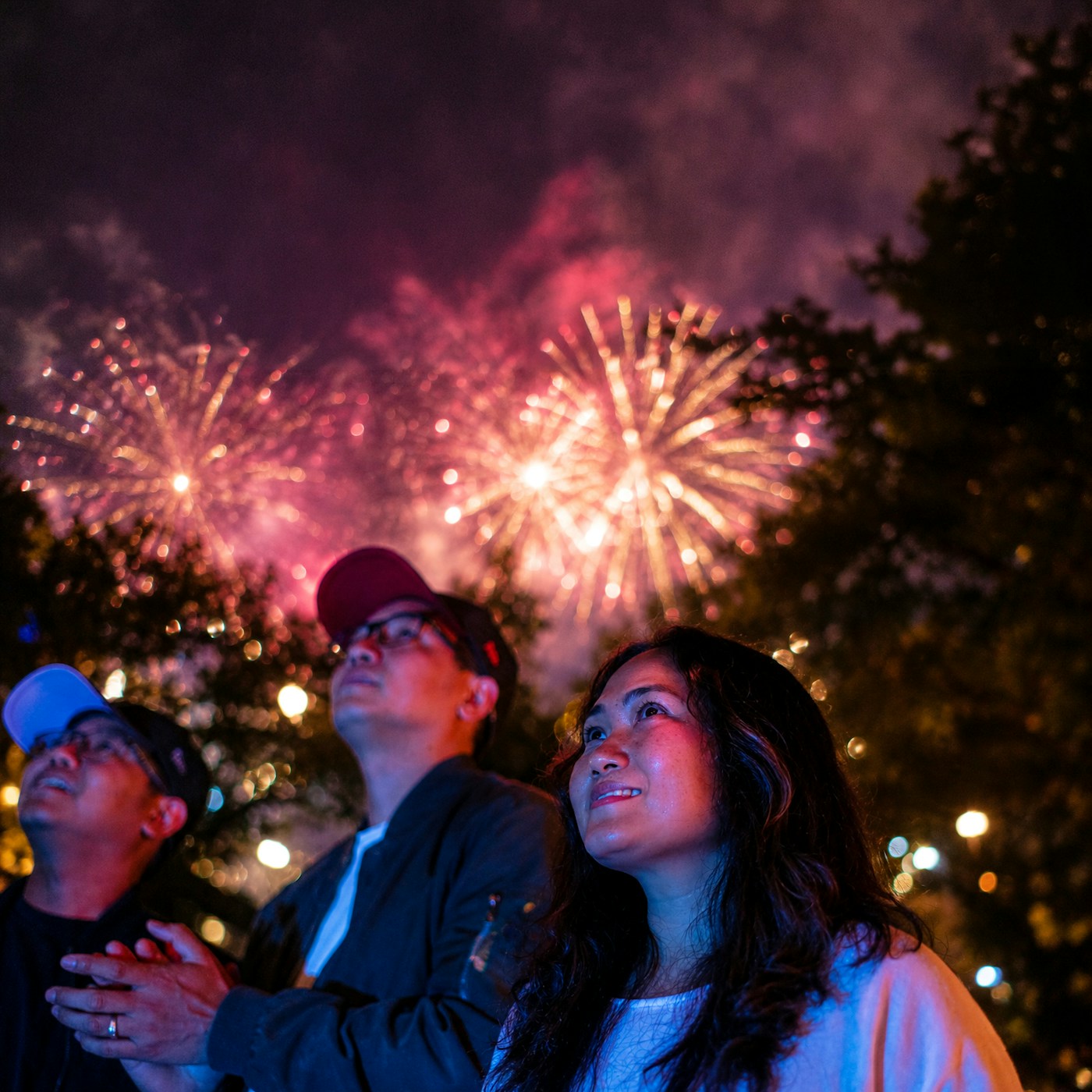 Three people watch colorful fireworks in the night sky, surrounded by trees and illuminated by bright bursts of light.
