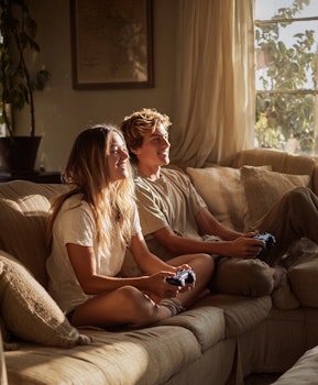 Two young gamers smiling and playing a console video game together on a sofa in a naturally lit living room