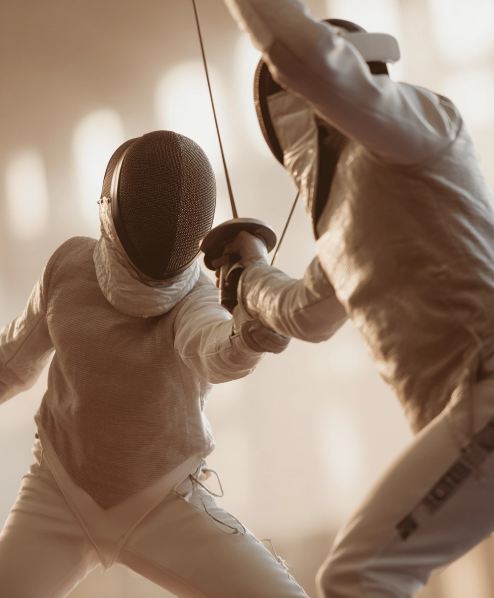 Two fencers in white protective gear mid-bout with crossed swords in a warmly lit training hall