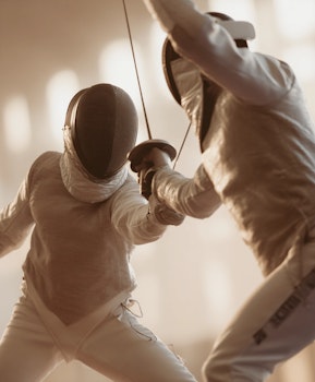 Two fencers in white protective gear mid-bout with crossed swords in a warmly lit training hall