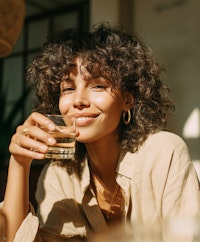 Woman with curly hair smiling while holding a glass of water in warm natural sunlight