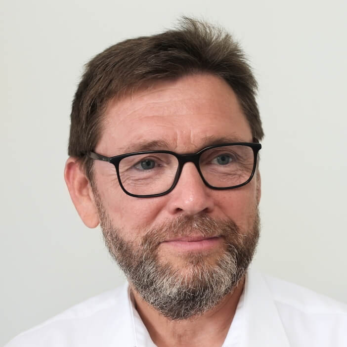 Close-up portrait of a middle-aged man wearing glasses and a white shirt, smiling slightly against a light background