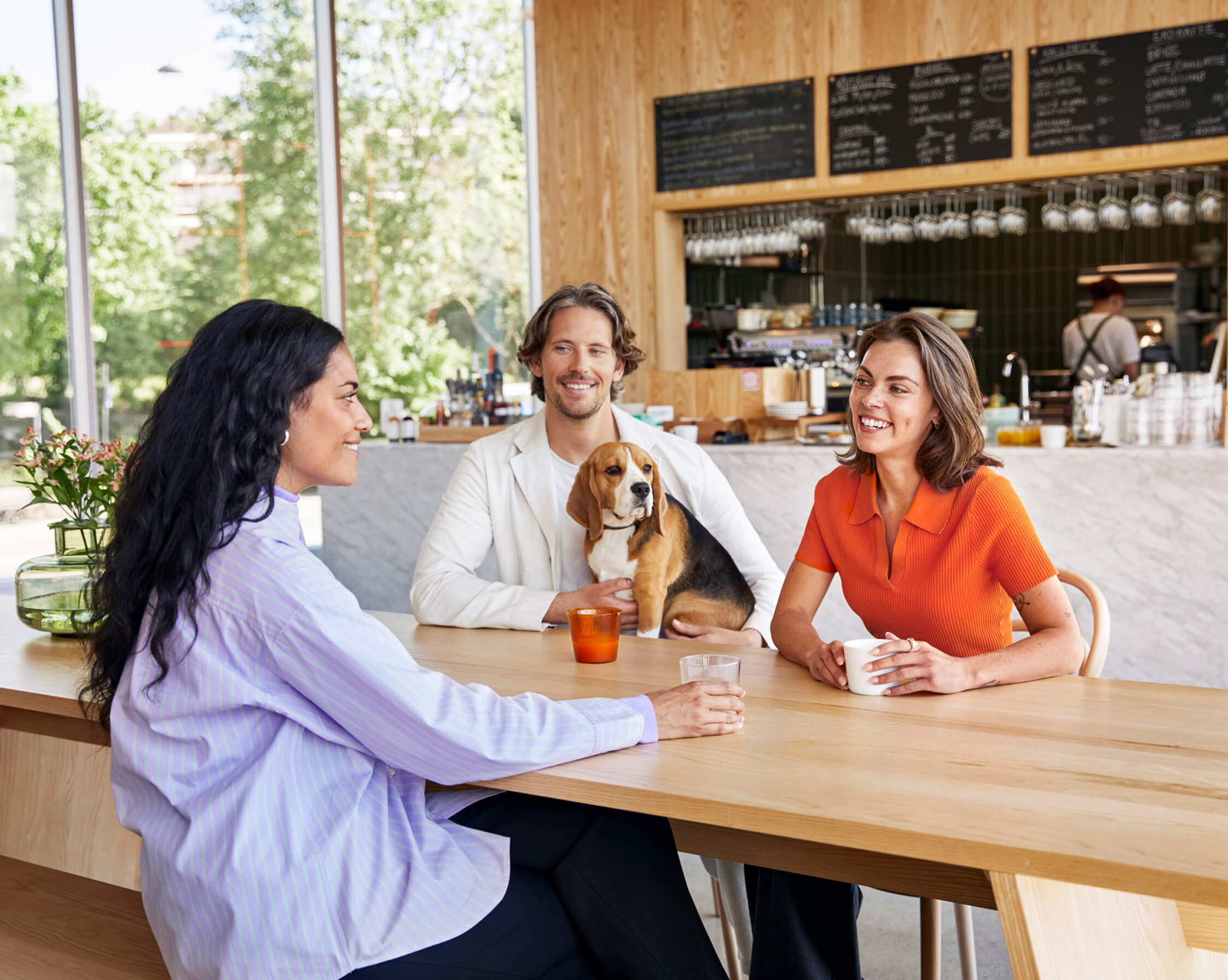 Three people sitting at a wooden table in a bright café, smiling and talking over coffee with a relaxed atmosphere