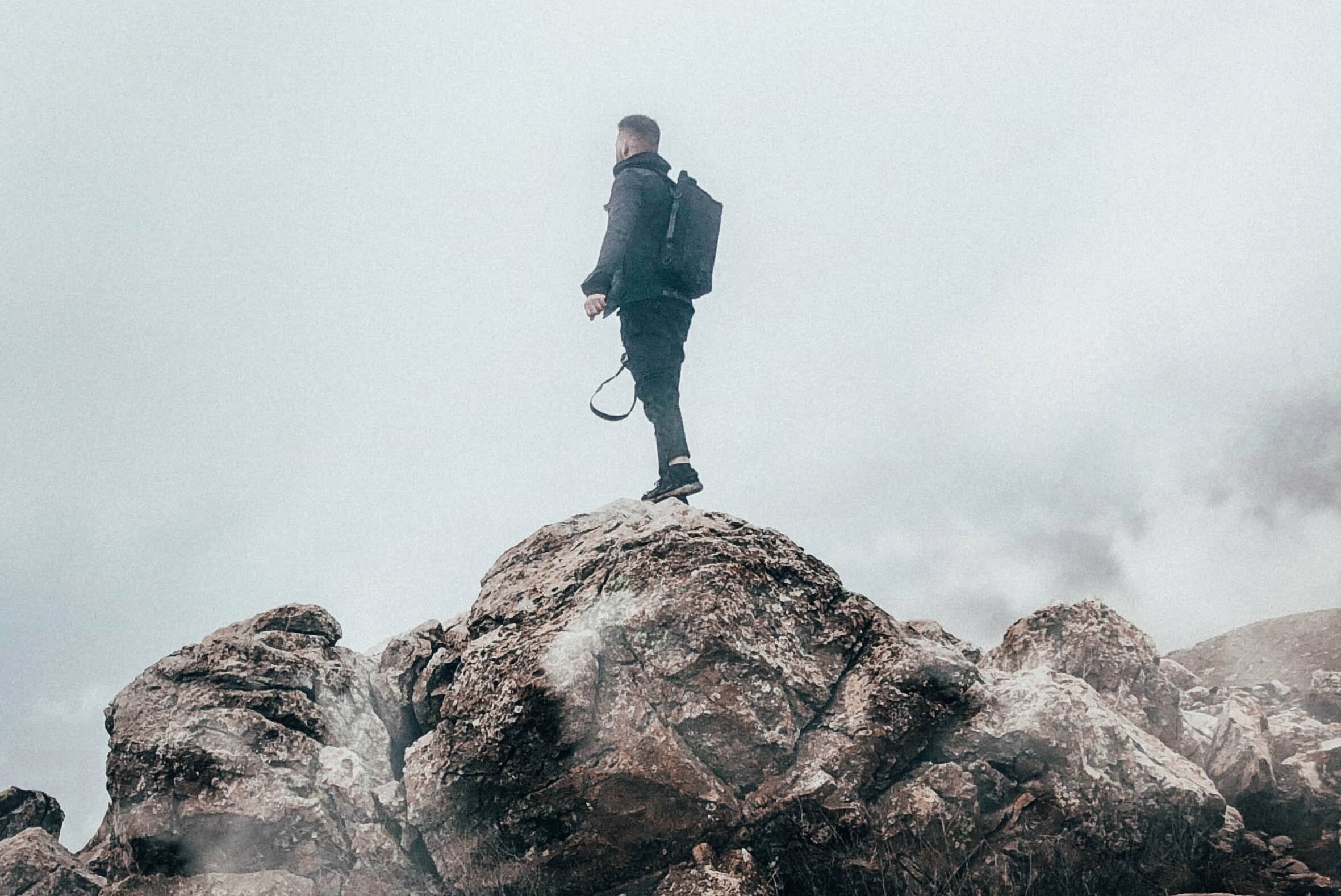 Person with a backpack standing on a rocky mountain peak, surrounded by fog and mist, looking into the distance
