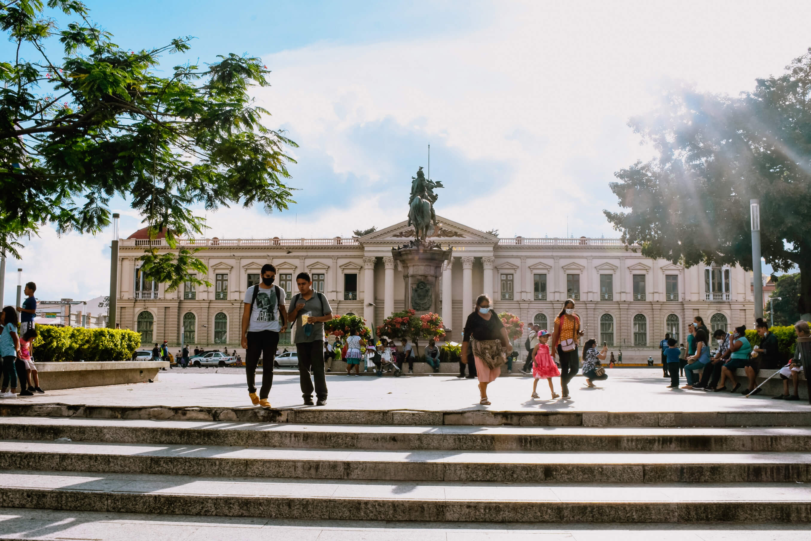 Busy public square in front of a historic building with people walking and sitting, featuring an equestrian statue in the center