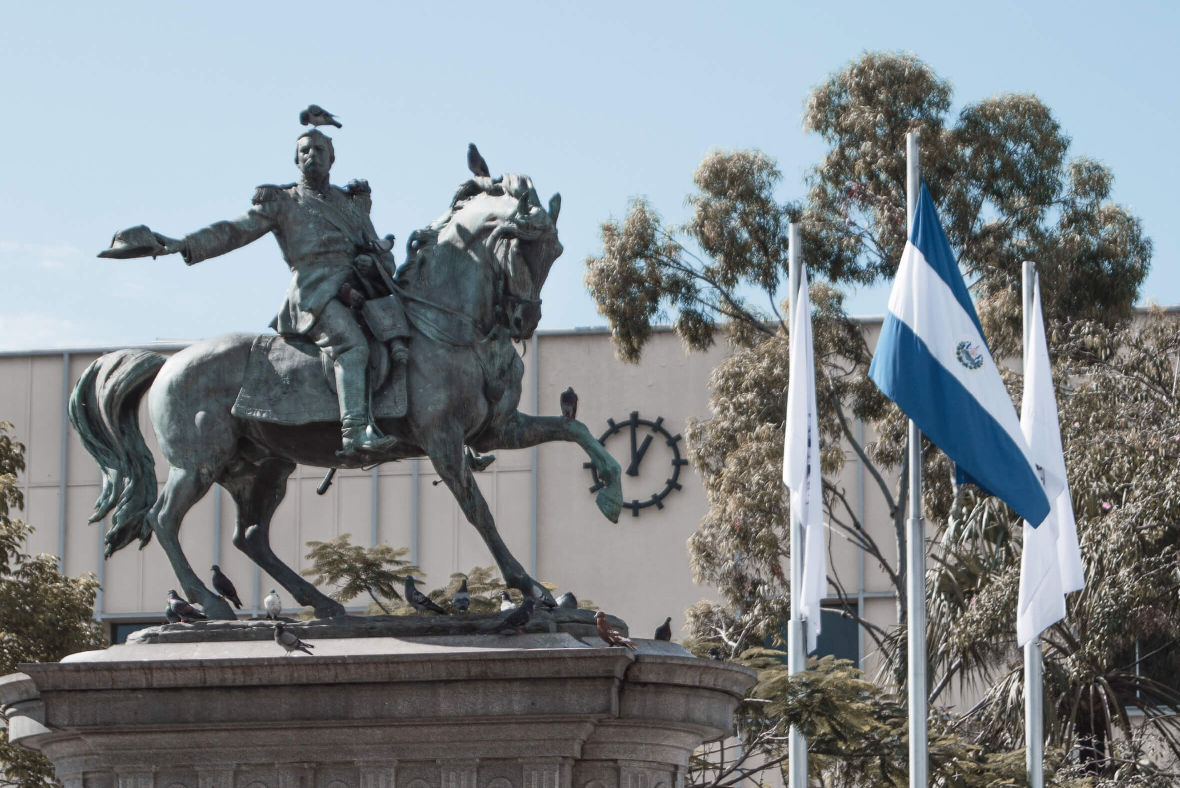 Equestrian statue of a man holding his arm out, with the flag of El Salvador waving beside it and trees in the background