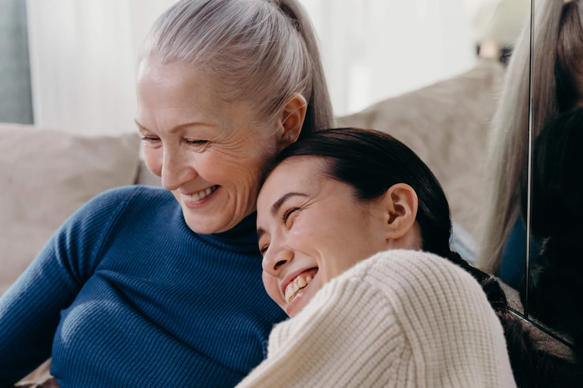 Older woman and younger woman sitting close together on a couch, smiling and laughing warmly in a cozy atmosphere