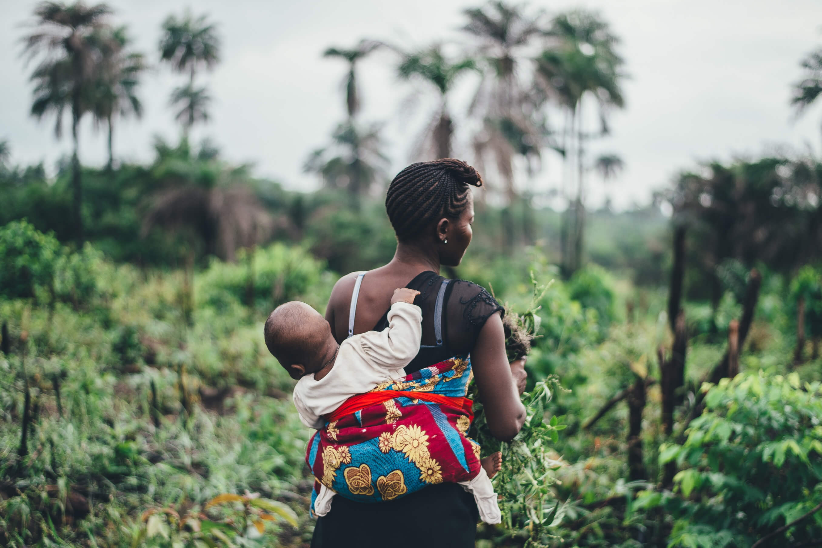 Woman seen from behind walking through a lush green field with a baby secured on her back using a colorful fabric wrap