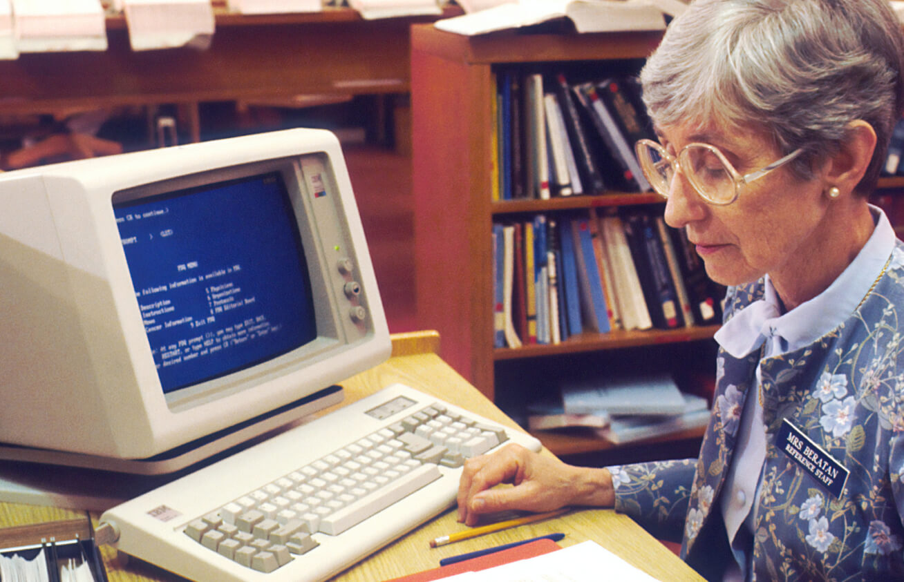Elderly woman sitting at a desk using a vintage computer with a blue screen in a library surrounded by books