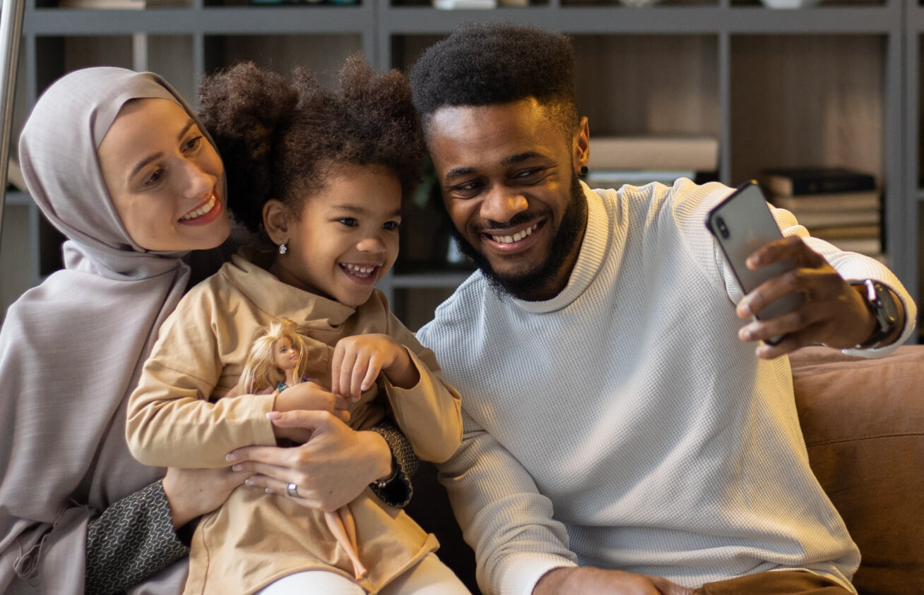 Happy family sitting on a couch taking a selfie together, with a young girl holding a doll and smiling at the camera