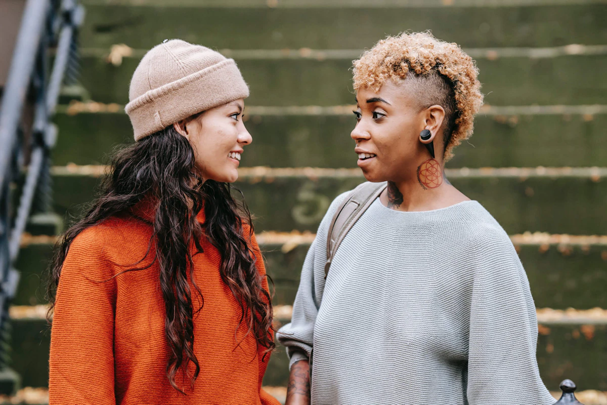 Two young women standing on outdoor stairs, wearing cozy sweaters, smiling and chatting with each other