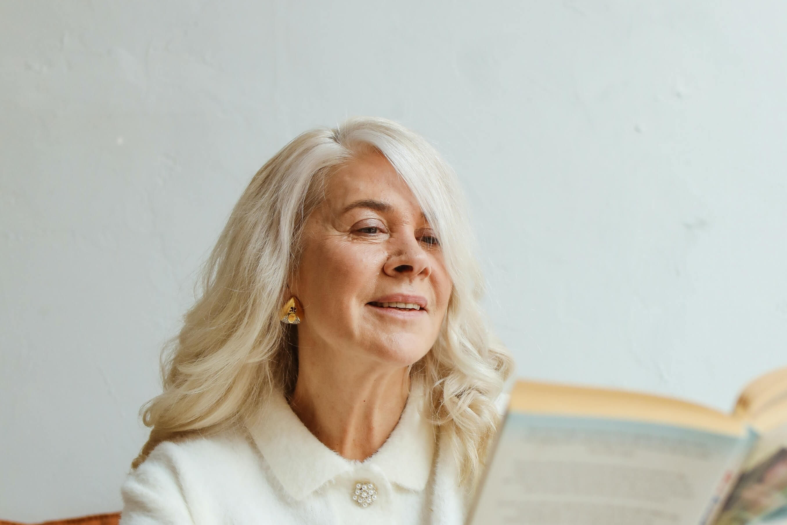 Elegant elderly woman with long blonde hair and white blouse reading a book and smiling softly