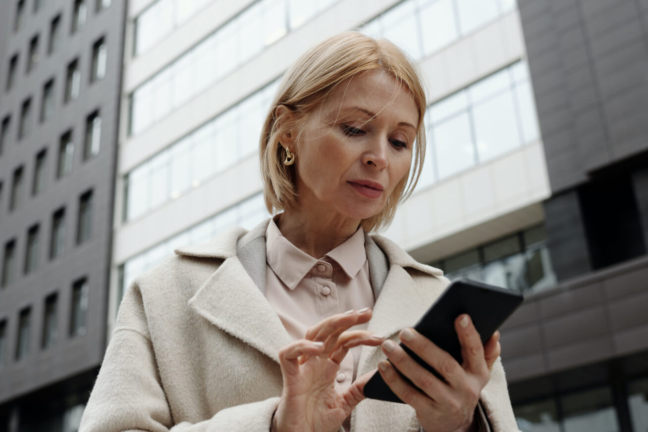 Woman with blonde hair standing in front of a modern office building, using her smartphone focused