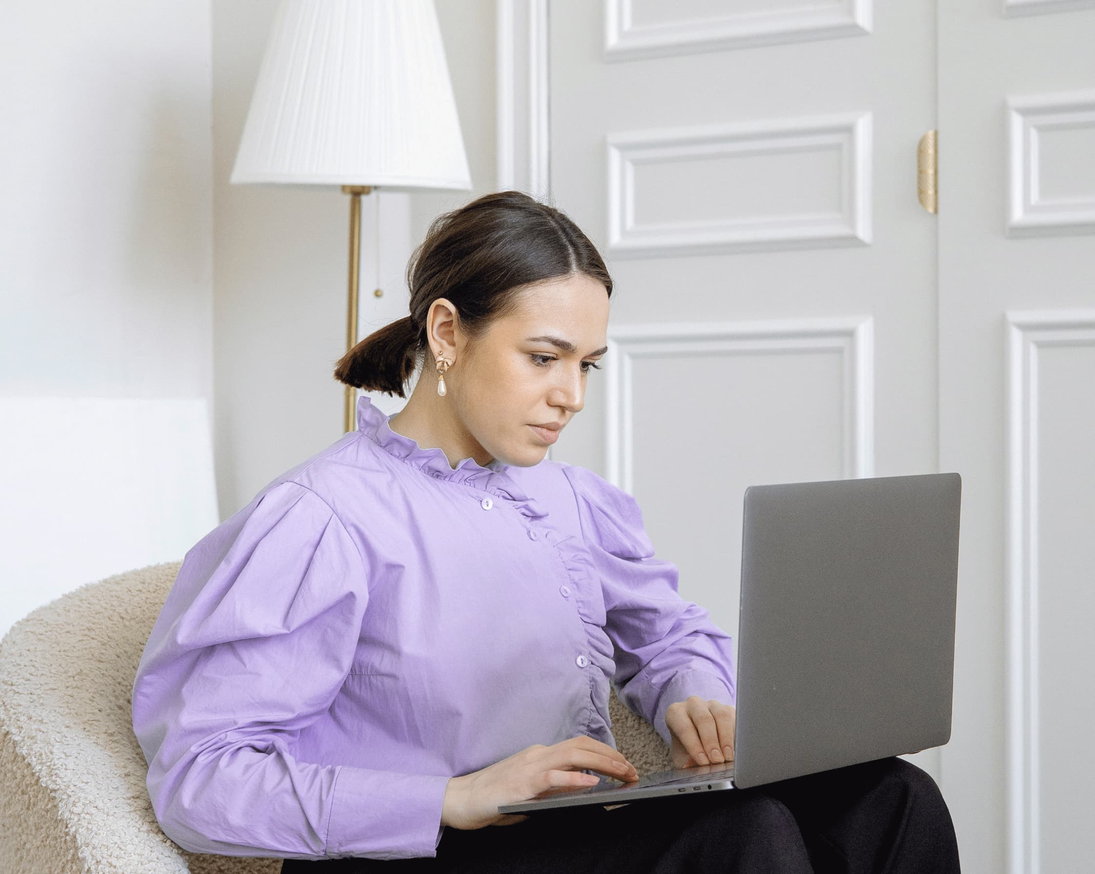 Woman in purple blouse sitting on a chair, working focused on a laptop in a bright room