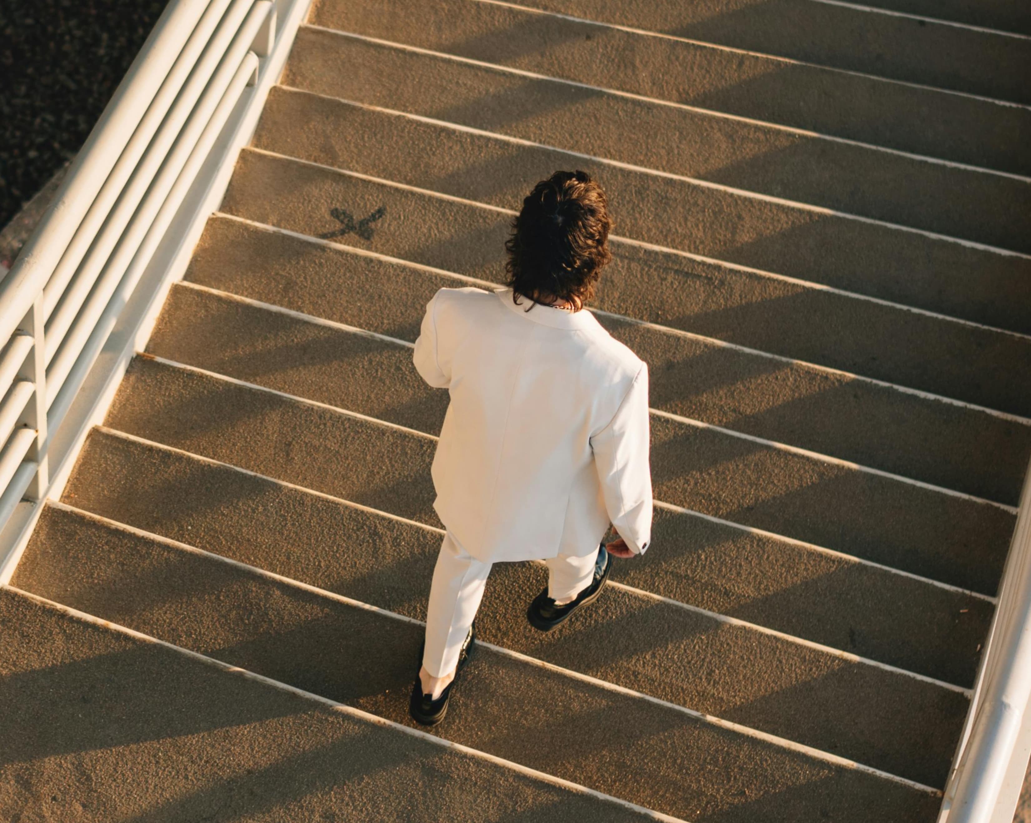 Man in a white suit walking down a wide outdoor staircase in sunlight, modern architecture in background