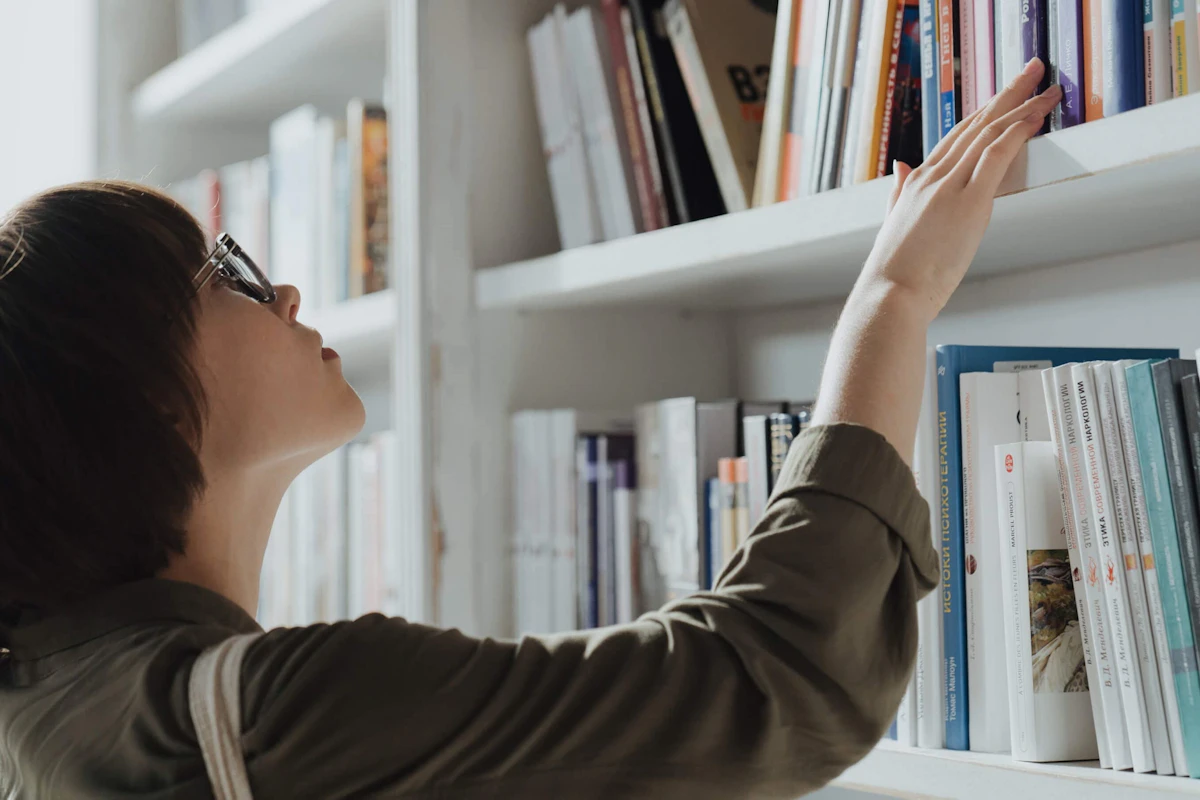 Woman with glasses standing in front of a bookshelf, reaching for a book in a bright modern room