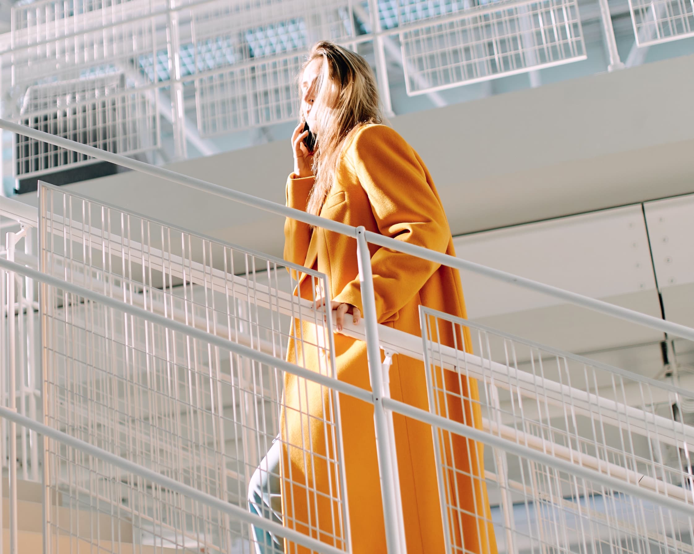 Woman in a long yellow coat standing on modern stairs, talking on the phone in daylight