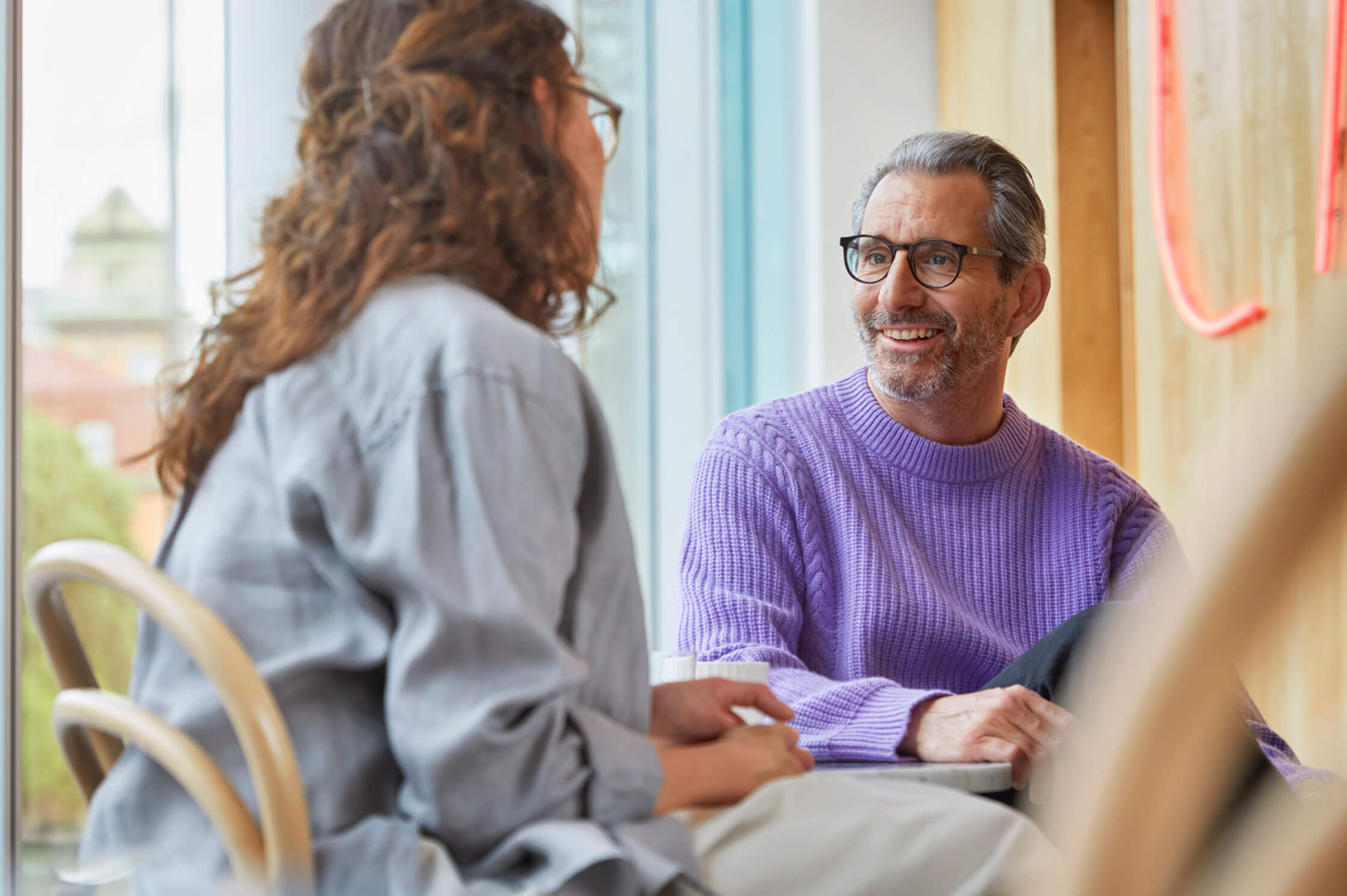 Man wearing a purple sweater and glasses smiling while talking to a woman in a café, natural light coming through windows