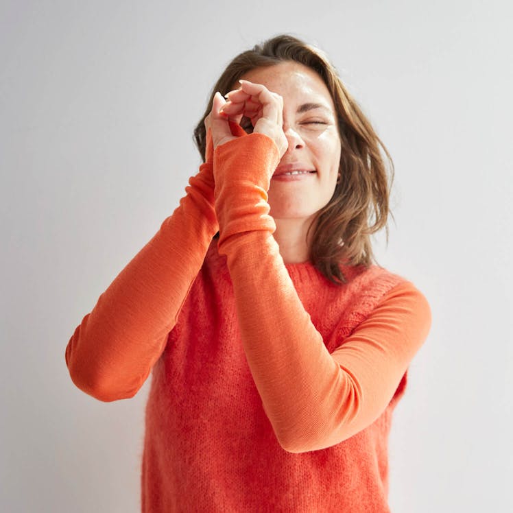 Woman wearing an orange sweater smiling playfully while making a gesture with her hands, standing against a light background