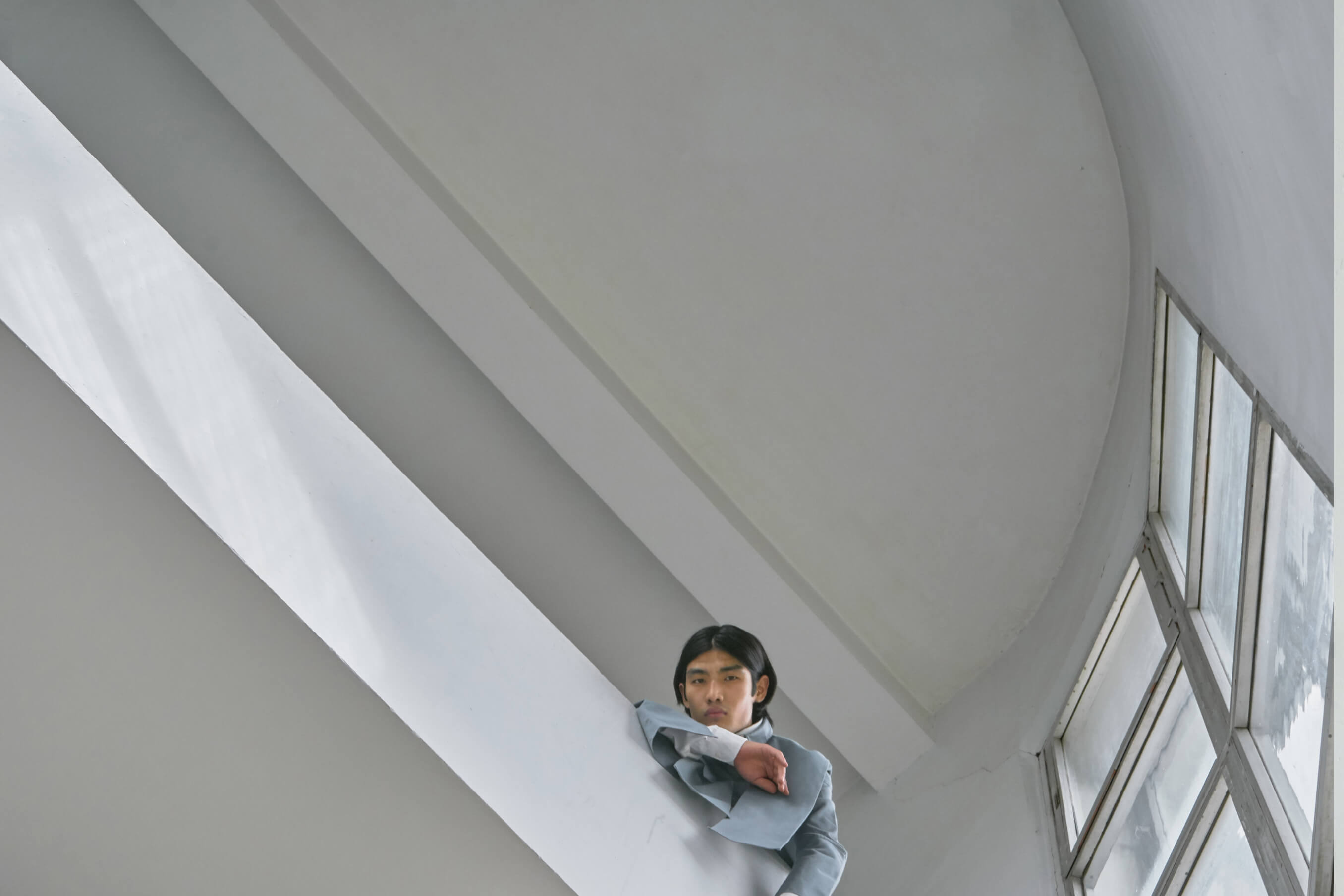 Man in grey suit leaning against a modern white wall with large windows, looking downward