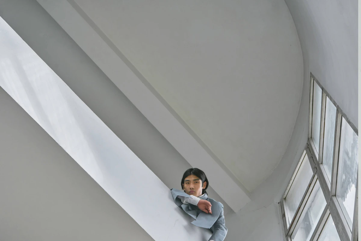 Man in grey suit leaning against a modern white wall with large windows, looking downward