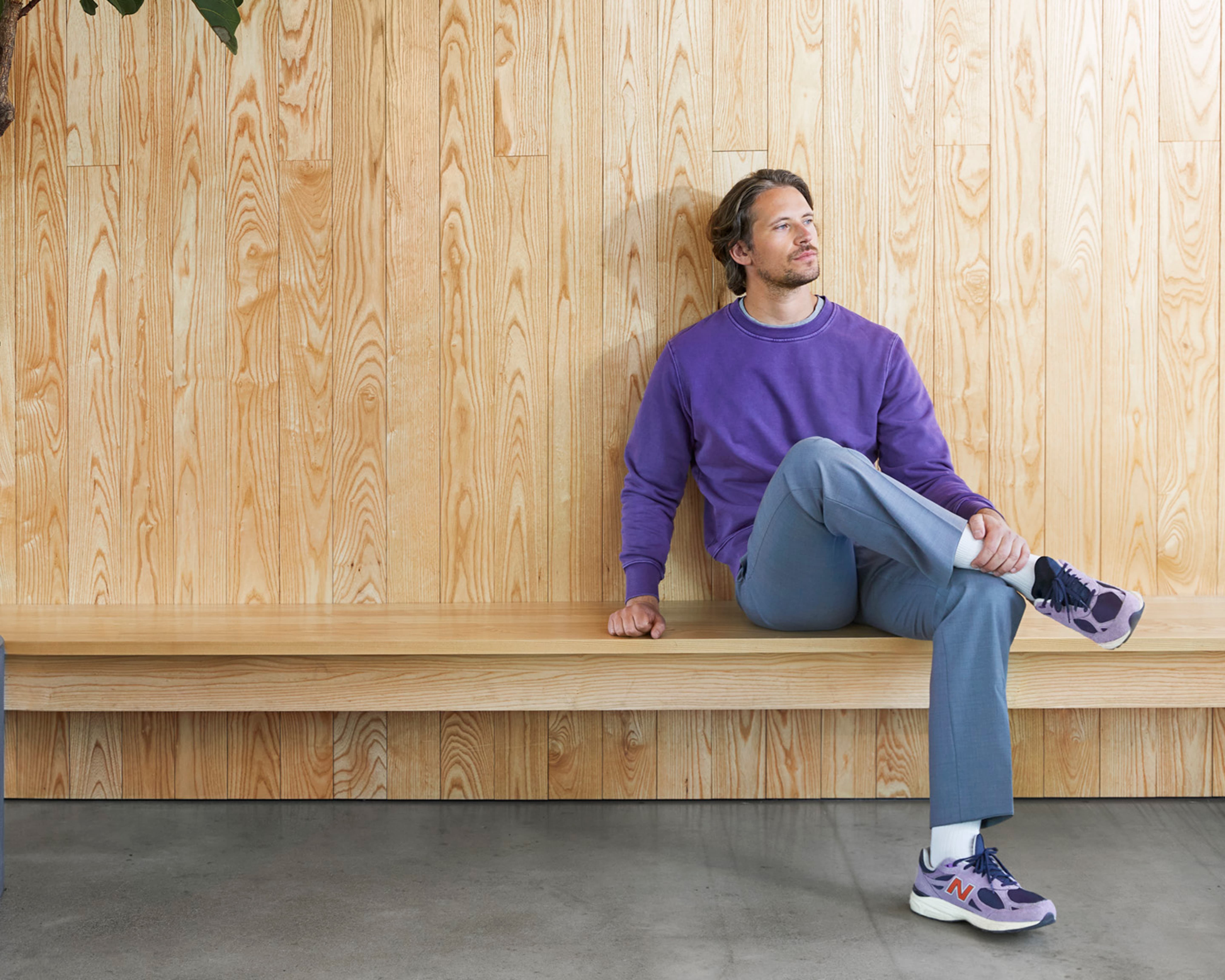 Man wearing a purple sweater and blue trousers sitting on a long wooden bench against a wooden wall, looking thoughtful