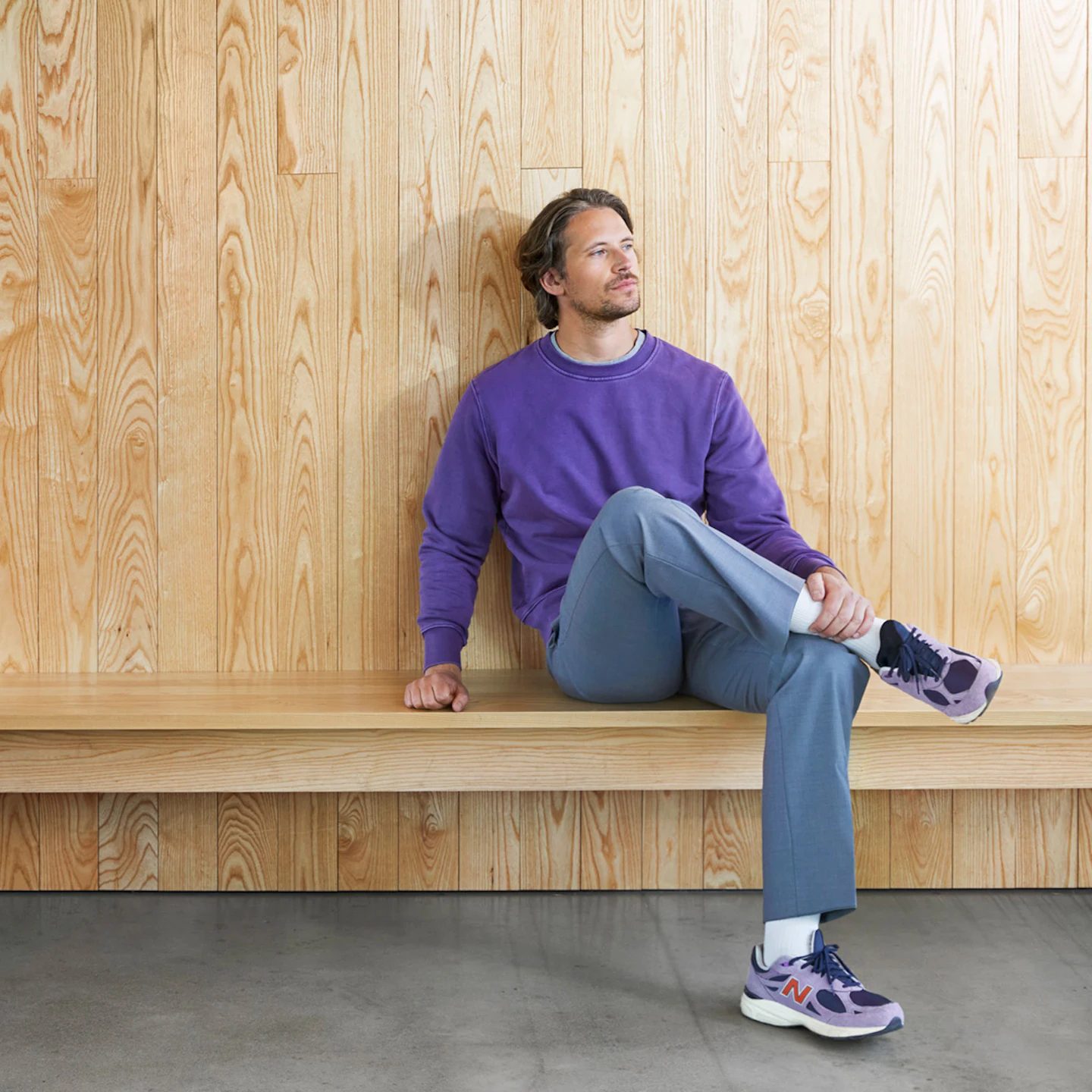 Man wearing a purple sweater and blue trousers sitting on a long wooden bench against a wooden wall, looking thoughtful