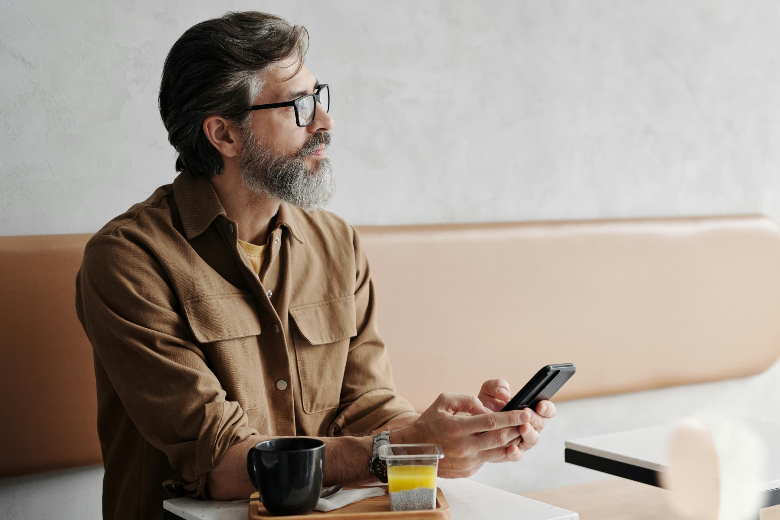 Man with glasses and beard sitting in a café, holding a smartphone with coffee and juice on table