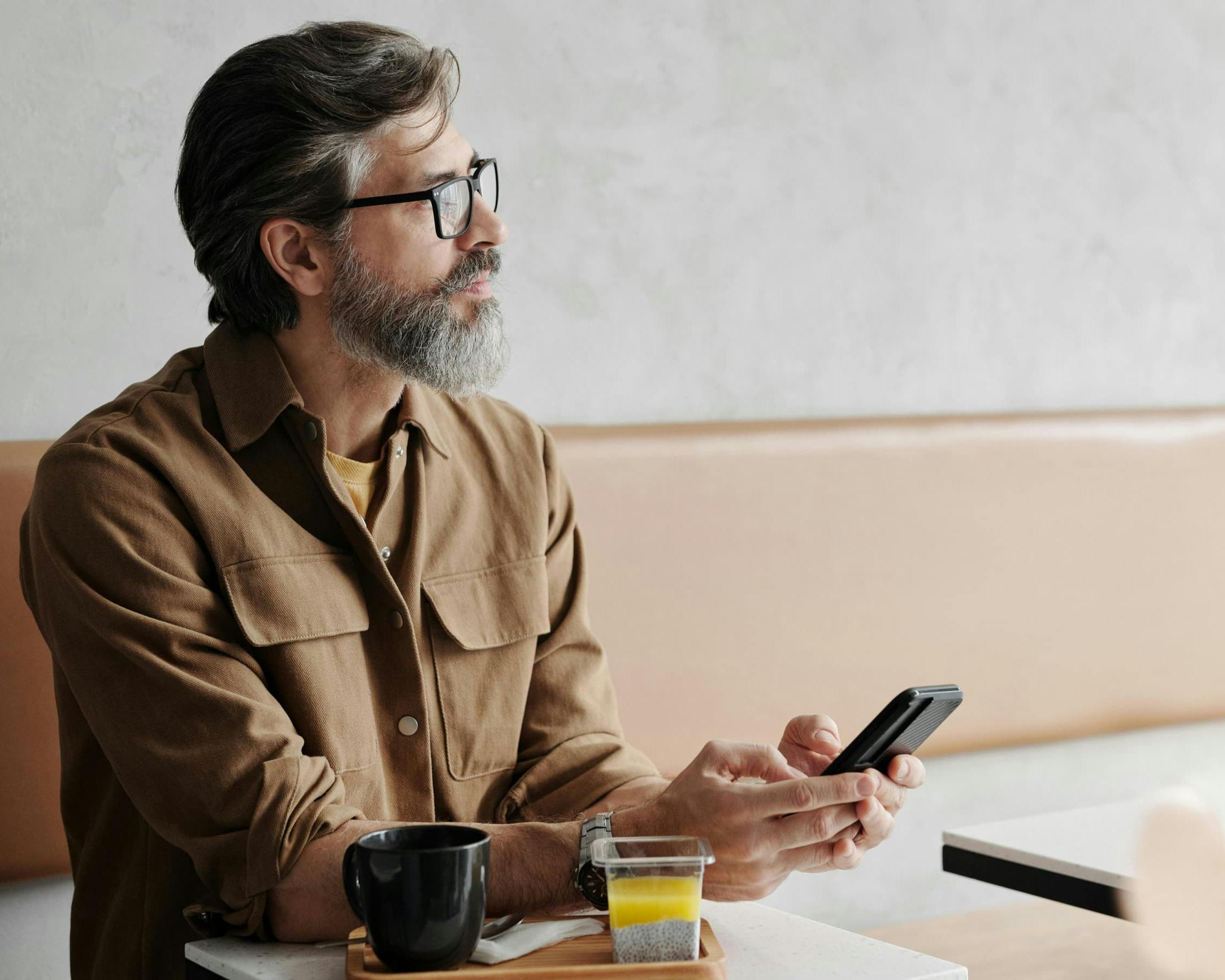 Man with glasses and beard sitting in a café, holding a smartphone with coffee and juice on table