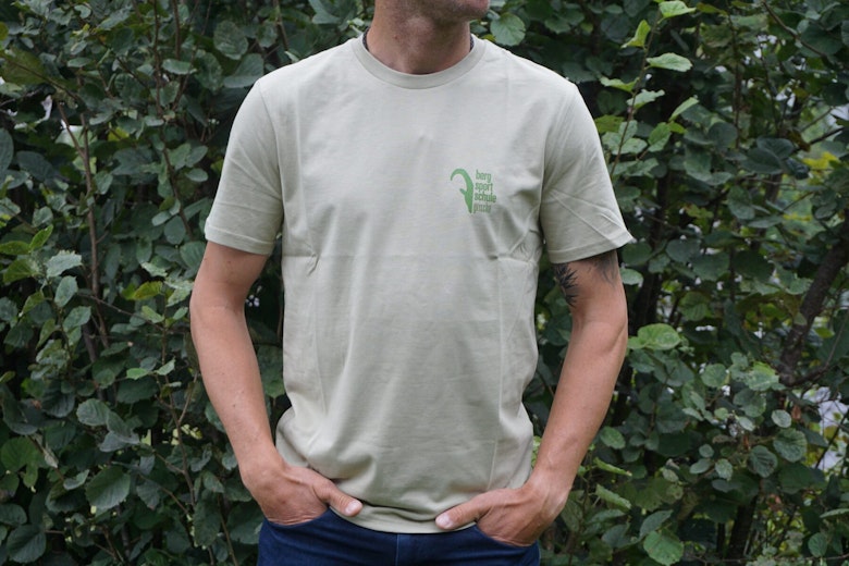 Eine Person in einem hellbeigen T-Shirt mit einem kleinen grünen Text und einer Grafik auf der Brust steht draussen in einem Gebüsch.