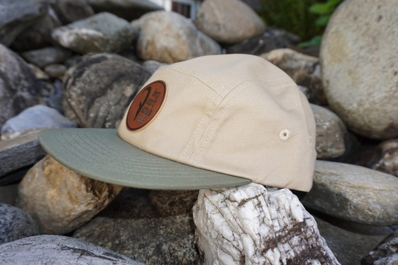 Eine beige-grüne Baseballkappe mit einem braunen Aufnäher sitzt aussen auf Steinen.