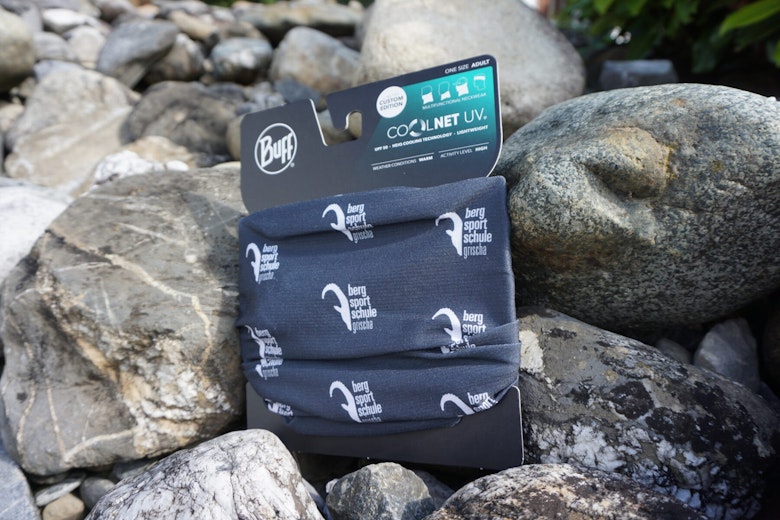 Eine Halskrause der Marke Buff mit dem Schriftzug "berg sport schule grindelwald" ist auf felsigem Outdoor-Boden abgebildet.