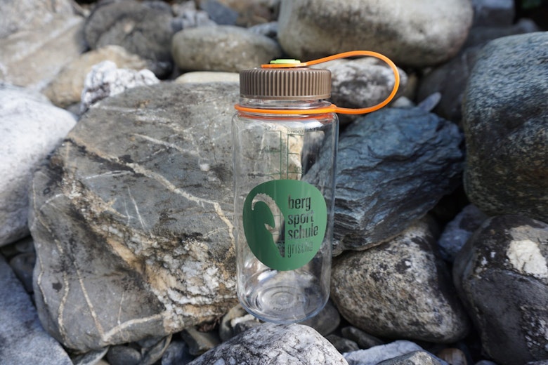 Klare Plastikwasserflasche mit braunem Deckel und orangefarbenem Band auf graüm Felsen; grünes Logo mit der Aufschrift "berg sport schule".