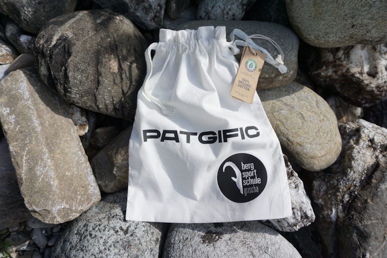 Ein weisser Beutel mit Kordelzug und der Aufschrift "PATGIFIC" und "berg sport schule" ruht auf grossen graün Steinen, an denen ein braunes Schildchen befestigt ist.