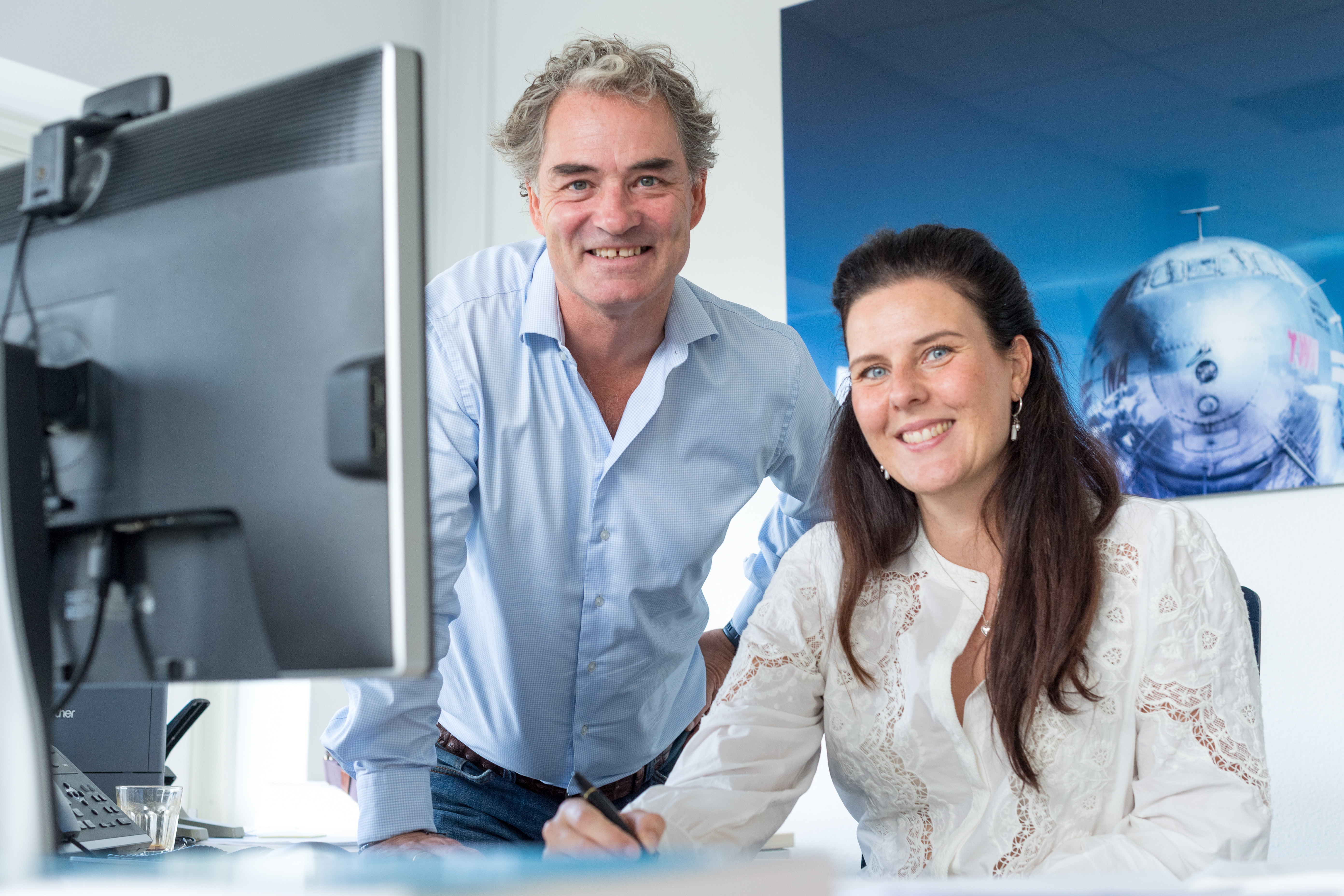 Twee personen achter een computerscherm, kijkend in de camera