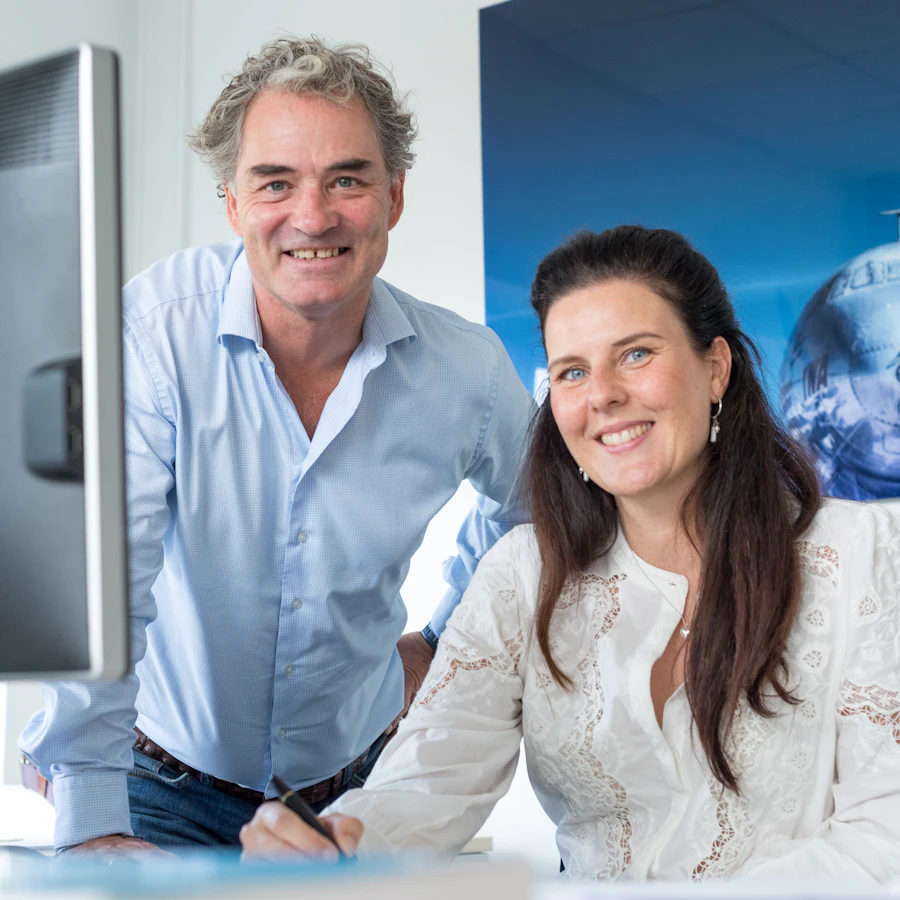 Twee personen achter een computerscherm, kijkend in de camera