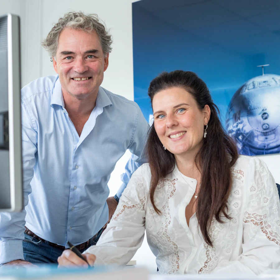 Twee personen achter een computerscherm, kijkend in de camera