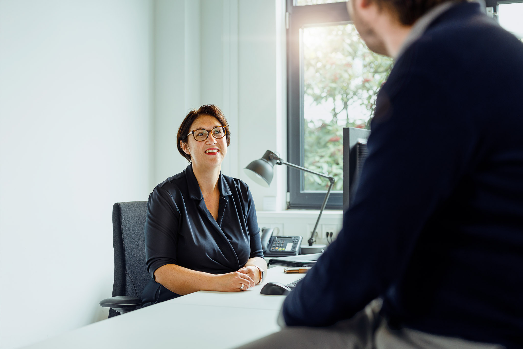 Twee personen die met elkaar in gesprek zijn. Eentje zit achter een bureau, de ander staat onscherp op de voorgrond.