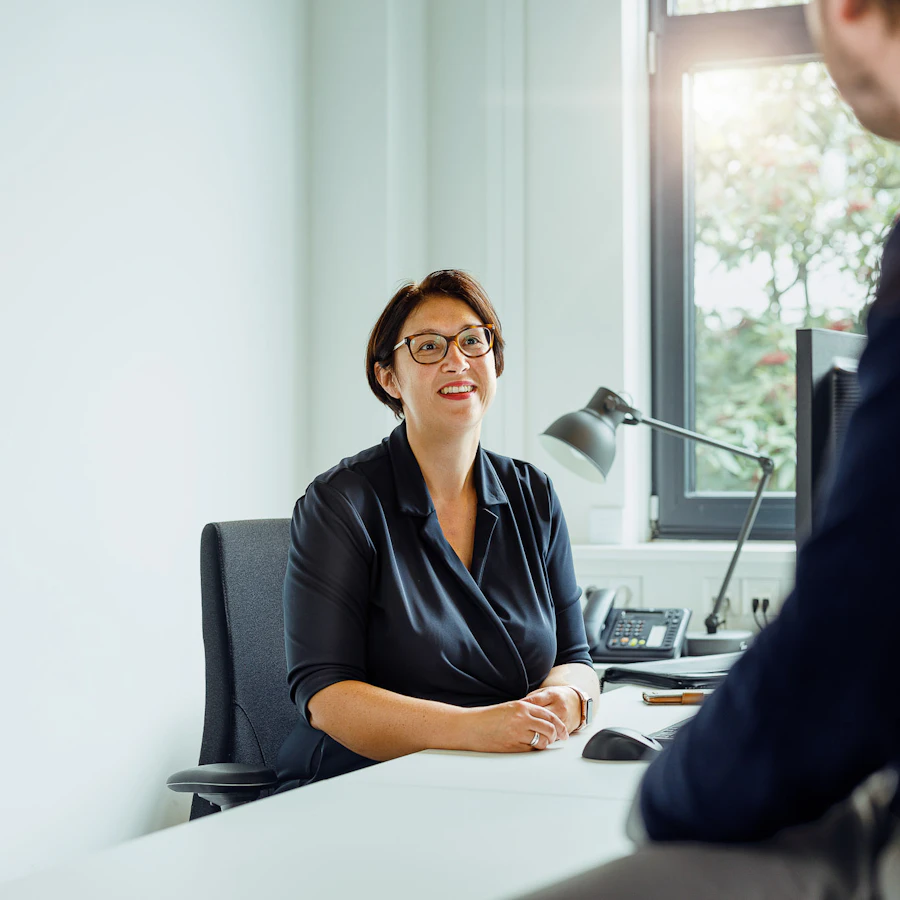 Twee personen die met elkaar in gesprek zijn. Eentje zit achter een bureau, de ander staat onscherp op de voorgrond.